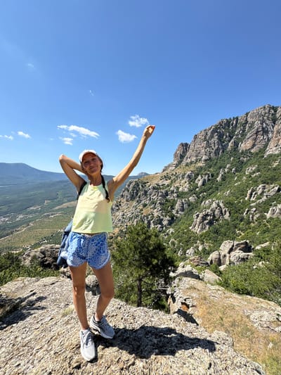 A young woman stands on a rocky outcrop in a mountainous area, with a panoramic view of a valley and distant mountains under a clear blue sky. She is wearing a white baseball cap, a light yellow tank top, blue and white patterned shorts, and grey athletic shoes. Her right arm is raised, with her hand holding a small, light-colored object, possibly food. Her left arm is bent, with her hand resting on the back of her head. She has braided hair and a slight smile. A backpack is slung over her left shoulder, and a folded blue and white checkered shirt is attached to her shorts. The terrain is rugged and rocky, with sparse green vegetation and trees scattered across the slopes. The background features distinct rock formations and a vast expanse of green hills and mountains, indicative of a natural, undeveloped landscape. The specific location is the Valley of Ghosts, near Luchistoe, Ukraine.