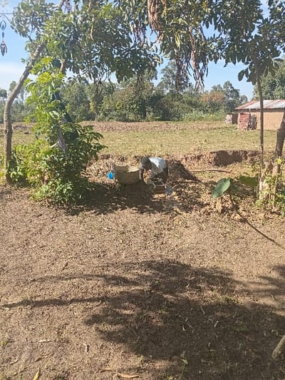 A rural landscape displays sparse vegetation and exposed dirt. A building rests in the background, nestled among trees. Foreground shadows partially obscure the terrain. A concrete structure with pipes sits prominently in the center, and miscellaneous items are scattered near it. Trees are visible on the left. The sky is light and clear.