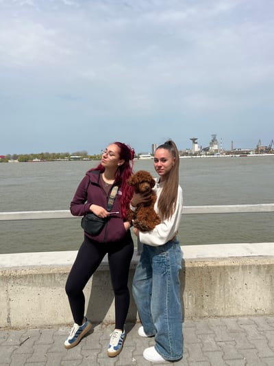 Two women are standing on a concrete ledge near a body of water in Nikopol, Bulgaria. One woman has long brown hair pulled back in a ponytail, and she is wearing a white sweater and blue jeans. She is holding a small, brown poodle dog. The other woman has long red hair and glasses. She is wearing a maroon jacket, black leggings, and sneakers with blue stripes. She has a black fanny pack strapped across her chest. The water stretches out behind them, and in the distance, there are industrial buildings and cranes. The sky is overcast. The pavement is composed of interlocking gray bricks.