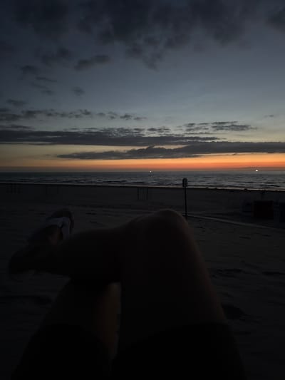 A first-person perspective shot of a person sitting on a beach at dusk. The person's legs are visible in the foreground, silhouetted against the darker sand. They are wearing white flip-flops. In the midground, the beach stretches out, with a few dark structures resembling beach chairs and a signpost. The sea is visible in the distance, with gentle waves. The horizon is marked by a band of orange and yellow light, indicative of a sunset or sunrise. Above, the sky transitions from a deep blue at the top to lighter shades of grey and blue closer to the horizon, with scattered dark clouds. The overall scene conveys a sense of peaceful solitude at the end of the day. The location is the beach on the island of Langeoog, Germany.