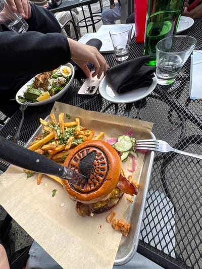 An outdoor dining scene shows a table set with a burger, fries, and a salad. A person's hand is reaching for a phone on the table. A knife is placed across the burger. In the background, a green bottle and red cup are on the table. The table is a metal mesh surface, commonly found in outdoor dining areas. This scene is likely in Anchorage, Alaska, United States of America, given the presence of a black bear motif on the burger bun.