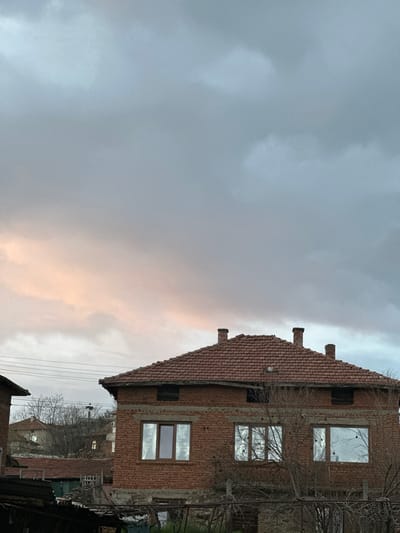 The image shows a brick building with a terracotta tile roof in Karnalovo, Bulgaria. The building has three visible windows reflecting the cloudy sky. There are bare branches in front of the building and power lines visible in the foreground. The sky is overcast with hints of pink light. Other buildings can be seen in the distance.