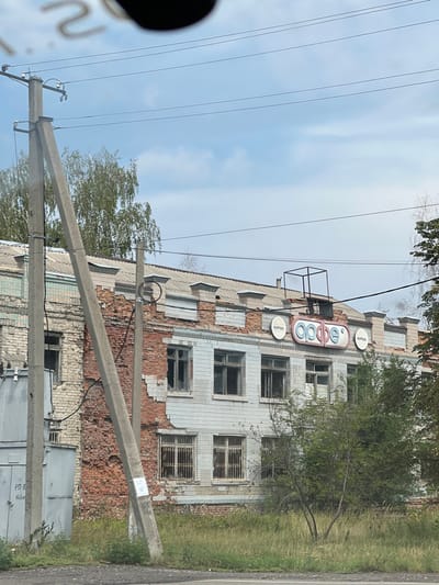 This image presents a two-story masonry building in Martove, Ukraine, showing significant damage. The left portion of the building reveals exposed red brick where the exterior cladding is absent, while the right portion retains light-colored square tiling with multiple missing sections. Numerous windows on both levels are broken or have missing panes, with the ground-floor windows on the right side secured by metal security grates. The building's upper facade features three circular signs: a central, larger sign displaying the Cyrillic word "орфе" (Orfei), flanked by two smaller signs, each bearing the Cyrillic word "ФИРМА" (FIRMA). A black, skeletal metal frame is positioned on the roof above the central sign. In the immediate foreground, a concrete utility pole with visible insulators and wires leans towards the building, partially obscuring a light-colored utility box with Cyrillic markings to the far left. The ground in front of the building is covered with overgrown grass and sparse small trees. The sky above is light blue with scattered white clouds. No people, active processes, or interactions are visible within the frame.