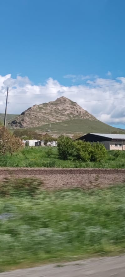 The image depicts a landscape in the vicinity of Spitak, Armenia, on a partly cloudy day. In the background, there is a pyramidal mountain with rocky patches and green vegetation, possibly grazing land. In front of the mountain, there are a few low buildings, some possibly industrial or agricultural. Closer to the viewer, there is a field with green grass and possibly newly tilled earth. The blurry foreground suggests that the image was taken from a moving vehicle. There are power lines visible stretching across the frame.