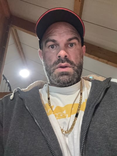 The scene is an indoor setting in Jemelle, Belgium. A single adult male is captured in a head-and-torso shot, looking directly into the camera. He possesses a dark beard and mustache. His expression is characterized by wide eyes and a slightly open mouth. He wears a black baseball cap with a red brim. His clothing includes a dark grey zip-up hooded jacket, lined with light-colored fleece visible at the collar, over a white t-shirt that displays a partial yellow graphic design on the chest. A prominent gold-colored chain necklace is worn over the t-shirt. The background features a sloped, light-colored ceiling with exposed wooden support beams. A bright, circular light fixture is visible in the upper-left quadrant. A curved, dark-colored architectural element, consistent with a spiral staircase railing, is partially visible in the lower-left. A small, light blue object or reflection is discernible in the upper right background.