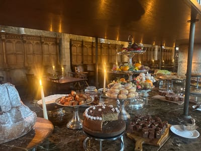 The image displays a meticulously arranged dessert and fruit buffet on a dark, marbled table within a large, dimly lit hall. The architectural style suggests an older building, possibly a banquet hall or chateau, consistent with venues found in Cernay-la-Ville, France. The walls feature extensive, ornate dark wood paneling in the upper sections, with a lighter-toned base of stone or plaster. Warm ambient light emanates primarily from numerous lit white taper candles positioned on the buffet table and on distant tables in the background.
No individuals are present in the scene.
The buffet, organized on silver platters, tiered stands, and wooden boards, includes:
- A large, light-colored baked good, possibly a Kugelhopf or Bundt cake, dusted with powdered sugar, situated on a wooden serving board on the left.
- A silver platter holding numerous golden-brown pastries or small baked goods.
- Multiple silver-tiered stands laden with fresh fruits, including pomegranates, apples, oranges, and green and red grapes. One lower tier prominently displays a large pile of powdered sugar-dusted donuts.
- A round cake, light in color with a darker frosting and white decorative elements, from which one slice has been removed, resting on a small wooden cake stand.
- A wooden board displaying numerous square chocolate brownies or fudge pieces, some topped with pecans or nuts.
- Additional cakes, tarts, and pastries, including a green-topped cake and a dark chocolate cake with white frosting swirls, are visible on various stands and platters in the middle and right background of the buffet.
- A silver serving utensil rests on a small white saucer on the far right of the table.
The background shows additional wooden tables and chairs, sparsely arranged, further illuminated by smaller candles, indicating a dining area. The overall scene suggests a prepared but currently unattended feast or gathering.