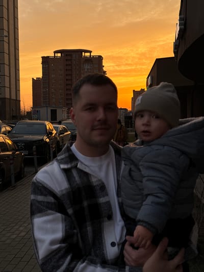 An adult male holds an infant outdoors in Gribovo, Russia, during an eventide or dawn with a prominent orange and yellow sky. The adult, positioned in the left foreground, has short dark hair and a neutral expression, wearing a white t-shirt under a black and white plaid shirt. He supports an infant or toddler, dressed in a grey beanie and a grey puffer jacket, who is also facing the camera with an open mouth. The immediate foreground features a dark grey paved sidewalk. A row of parked vehicles, separated by grey and white bollards, occupies the mid-ground to the left, including a dark SUV with a Russian license plate (P550). In the background, multiple multi-story residential buildings are silhouetted against the vibrant sky, one featuring a distinct rooftop structure. A "No Stopping" sign is visible on a pole near a small building in the mid-ground.