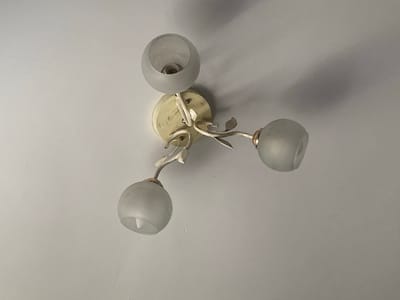 The image shows a close-up, overhead view of a ceiling light fixture. The fixture is cream-colored with three frosted glass globes, each housing a light bulb. The metal arms of the fixture are shaped like branches with small leaf accents. The ceiling is a plain, textured white. The scene is indoors, and there is no indication of the specific location in Keŋeş, Kyrgyzstan, other than it being a part of a building. There are no people or actions depicted.