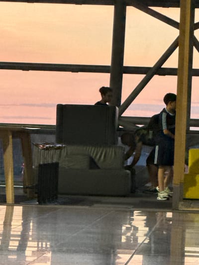 The image captures a scene at Brussels Airport in Zaventem, Belgium, during sunset or dawn. The sky displays a gradient of soft pink and orange hues. In the foreground, a polished floor reflects the surrounding structures and light. Several people are visible. On the right, a young male wearing a dark blue tracksuit with white stripes and white sneakers stands facing left. Next to him, a person is hunched over, appearing to be handling some equipment. To the left, a woman with dark hair tied up is partially visible behind a large, dark, rectangular object, possibly a piece of luggage or equipment. Another dark, rectangular object, potentially a suitcase, is positioned in front of her. Behind these individuals, a metal support structure, characteristic of an airport terminal, is evident. The overall atmosphere suggests a moment of transition or waiting within the airport environment.