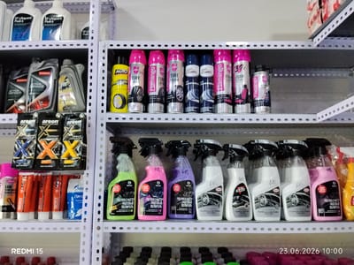 The image depicts a shelf in what appears to be an automotive supply store or a home garage. The shelf is filled with various car care products. On the top shelf, there are several white plastic bottles with blue labels that read "ZF Lifeguard Fluid 6". To the right of these are other bottles that are partially visible.

The middle shelf is more densely packed. On the left side, there are two bottles labeled "ACTIVE DEFENCE" and a bottle of "ZYPOKO". Further to the right, there is a row of aerosol cans. The first few cans are yellow with "AUTO" branding, followed by several pink and blue cans labeled "Flamingo" with text indicating "TIRE SHINE" or "FOAM CLEANER". There are also dark blue cans with "AUTORAMA" branding.

The bottom shelf displays a variety of spray bottles and tubes. On the far left, there is a green spray bottle labeled "GRASS POLYROLE MATTE". To its right are pink and purple spray bottles with similar branding. Then there are white spray bottles with "UNTEC" and "Dr. Active" branding. There are also several orange tubes and a blue plastic bag with Russian text that appears to be "Gazpromneft".

The overall environment is one of organized retail display, with metal shelving units. The lighting suggests an indoor setting. The date stamp in the bottom right corner indicates "23.06.2026 10:00". There is no visible indication of the specific location within Yovon, Tajikistan, other than the presence of the store itself. There are no people or animals visible in the image.