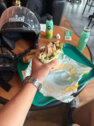 A person's arm and hand, wearing a silver watch, hold a partially eaten wrap over a green tray. The wrap appears to contain chicken, lettuce, and other ingredients. A black motorcycle helmet with a clear visor and the "PRUDEN" logo is visible to the left. Two green bottles, one with a black cap and a label indicating it's a "RAW" brand drink with a lemon graphic, and another open bottle, are on the table. A small white piece of paper and a QR code are also present. The scene takes place on a wooden table, suggesting an indoor setting like a cafe or restaurant. The environment is dimly lit. The location is identified as Samana bahu, India.