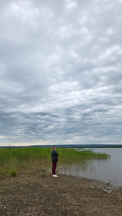 A person stands on the shore of a body of water, facing away from the camera. The person is wearing a dark jacket and dark red pants, with white shoes. They have their arms crossed. Behind the person is a thick growth of tall, green grass. The body of water appears to be a lake or slow-moving river, with the far shore visible in the distance. The sky is overcast with a pattern of layered, grey clouds. The ground in the foreground is composed of dirt and small pebbles. The scene is likely located in the village of Kupoyarovo, Russia, given the natural landscape.