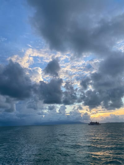 The image shows a seascape in Thong Nian, Thailand. A ferry is on the water, silhouetted against the setting sun on the horizon. The sea is rippled with sunlight reflecting off the water's surface. Dramatic clouds fill the sky, ranging from dark gray to bright white. Patches of blue sky are visible through the cloud cover.