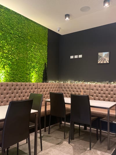 This image depicts the interior of what appears to be a modern restaurant, cafe, or lounge in Sondrio, Italy. The room is empty of people, with a focus on its contemporary decor and seating arrangements.
On the left side of the frame, a vibrant, bright green wall covered in artificial moss or a similar plant material dominates the lower half, illuminated from below to create a glowing effect. Above this, the wall transitions to a dark grey or black, and further up, a white perforated ceiling with integrated spotlights is visible. A small security camera is mounted where the white ceiling meets the dark wall.
The back wall is dark grey or black, featuring a single framed picture hanging centrally; it appears to be the iconic black and white "Abbey Road" album cover by The Beatles. Below the picture, three white electrical outlets or switches are vertically stacked on the wall. Along the top edge of a long, tufted banquette that runs parallel to the back wall, delicate string lights or fairy lights are draped, adding a warm, decorative touch.
To the right, several tables with white tops and dark bases are arranged in front of the banquette. Black chairs with sleek designs and dark frames are pulled up to these tables. The floor is a light grey, possibly concrete or tiled, providing a neutral base for the furniture.
The overall atmosphere is clean and modern, characterized by contrasting textures and colors, including the plush brown banquette, the organic green moss wall, and the stark black and white elements. The interior lighting, including spotlights and decorative string lights, suggests an indoor setting, potentially during the evening. No specific activity is taking place, and there is no visible text to read within the image.
