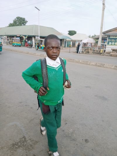 A young person in a green uniform, complete with a backpack, stands in the middle of a road. The person gazes directly at the viewer. In the background, there is a street with buildings, possibly shops or stores, visible along the roadside. The photo appears to be taken during daylight hours.