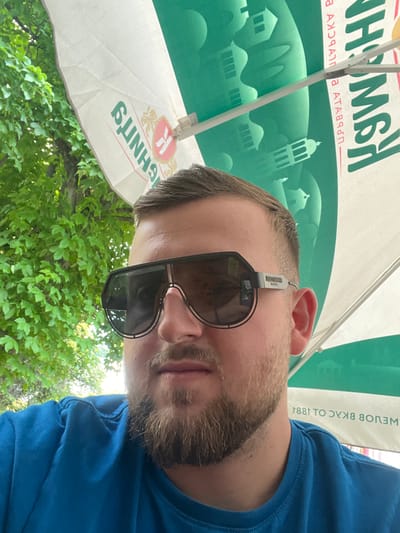 A man with a beard and sunglasses is seen from a low angle, possibly taking a selfie. He is wearing a blue t-shirt. Above him is a white umbrella with green writing and designs, as well as a depiction of a building resembling a church or tower. Behind the umbrella, green trees and foliage are visible. The writing on the umbrella includes "Kambana" and other text in Cyrillic. Given the text on the umbrella and the request to include the location of Sadovo, Bulgaria, it is probable that the photo was taken there.