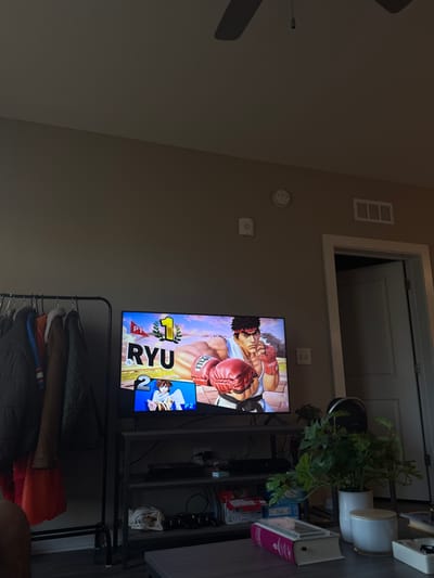 This image depicts an indoor living space, identified as being located in Gainesville, United States of America. The primary focal point is a large flat-screen television displaying a video game, "Super Smash Bros. Ultimate." The screen shows "P1 RYU" with a gold "1" signifying a round win, positioned above "P2" with an anime-style character, indicating "2" stocks remaining.

Below the television, a multi-tiered, dark-colored media console is laden with electronic devices, including various gaming consoles, controllers, and tangled wiring. To the left of the console, a black metal garment rack holds multiple coats and jackets in colors such as dark green, brown, and orange.

In the foreground, a dark coffee table supports a potted green houseplant, a white cylindrical container with a lid, a decorative box shaped like a book, and a small metal dish. The lower left corner of the frame shows a portion of a bare leg, suggesting a person is seated out of view.

The walls are painted a uniform light beige. Mounted on the wall are a white circular smoke detector, a white square wall panel (possibly a thermostat or intercom), and a rectangular white HVAC vent with horizontal louvers. To the right of the television, a white paneled door is partially open, revealing a darker interior space. A white light switch is visible on the wall adjacent to the door frame. A dark ceiling fan is partially visible at the top of the frame. The room is moderately illuminated by ambient light.