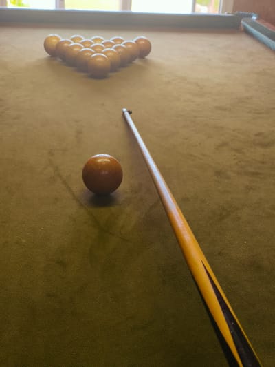 The image presents a high-angle view of a pool table situated indoors in Терновский округ, Ukraine. The table is covered in a textured, olive-green felt. Fifteen solid, light-brown pool balls are arranged in a triangular rack formation at the far end of the table. A single, larger, light-brown cue ball is positioned in the foreground, offset to the left of the central axis. A pool cue, predominantly yellow with dark triangular inlays along its lower shaft, extends diagonally across the felt from the bottom right to the middle left, its tip pointing towards the cue ball. The dark-colored rails of the pool table are visible along the upper perimeter of the frame. In the background, a window with white mullions is present, admitting diffused natural light into the room and revealing blurred outdoor greenery. No individuals are visible in the scene.