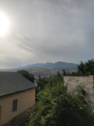 The image depicts a view of a distant mountain range under a cloudy sky, with the sun partially visible on the upper left. In the foreground, there is a light-colored building with a dark, corrugated roof, and a single window. To the right of this building is a partially visible brick structure. Lush green trees and vegetation are present in the midground, obscuring some of the view of the landscape and buildings. The overall scene suggests an outdoor environment, possibly on the outskirts of a town or city, with hints of residential or industrial structures blended with natural elements and a prominent mountain backdrop.