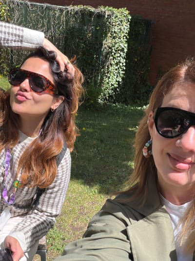 Two women are captured in a selfie outdoors on a sunny day. The woman on the left has long, wavy brown hair, is wearing large, tortoiseshell-patterned sunglasses, and a white and brown patterned sweater. Her hand is on her head, and she is puckering her lips. She is also wearing a lanyard with several colorful charms. The woman on the right has shoulder-length, auburn hair and is wearing large black sunglasses. She has hoop earrings with colorful beads and a nose ring. She is wearing a green jacket over a white t-shirt and is smiling. They are both seated on what appears to be a bench. The background consists of lush green grass, ivy-covered walls, and a brick wall, suggesting a garden or park setting. The location of Epsom and Ewell in the United Kingdom is not discernible from the visual elements in the image.