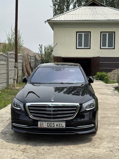 A black Mercedes-Benz S-Class sedan is parked on a concrete driveway in front of a two-story house. The house is painted yellow with a white corrugated roof and has two square windows on the upper floor. To the left of the house is a concrete fence and a wooden utility pole. The license plate on the car reads "01 001 KEL" with "KG" below the "01". The scene appears to be in a residential area in КРУГОВО, Kyrgyzstan, with trees and vegetation visible in the background. The sky is overcast.