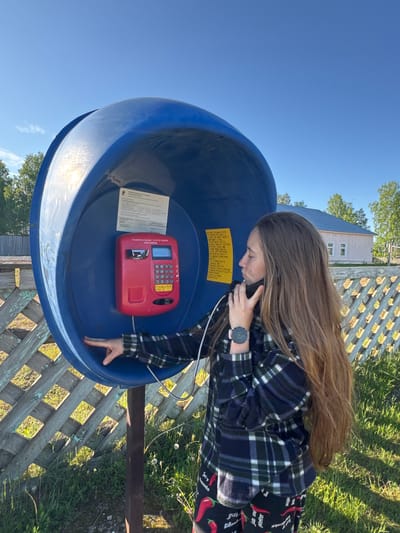 An outdoor scene in Ust'-Silayka, Russia, captures a young female interacting with a public telephone booth under a clear blue sky. A prominent blue, shell-shaped public telephone booth stands centrally, supported by a dark pole. Inside the booth, a red payphone with a digital display, keypad, and Braille markings is mounted on the rear wall. A white document with Cyrillic text is affixed above the payphone, and a yellow handwritten note is taped to its right.

A female with long, light brown hair, wearing a dark plaid long-sleeved shirt and dark pants with red chili pepper patterns, is positioned to the right of the booth. She holds the red payphone receiver to her right ear with her right hand, while her left arm extends into the booth, with her index finger pointing towards the lower-left interior. A light-colored smartwatch is visible on her left wrist.

In the background, a weathered wooden lattice fence with peeling light blue/green paint runs horizontally across the mid-ground. Beyond the fence, green grass and sparse vegetation are visible. To the right, a light pink house with a dark blue roof and two white-framed windows is partially visible behind the fence. Green trees are present in the far background on both the left and right sides of the frame. The scene is illuminated by bright sunlight, casting distinct shadows.