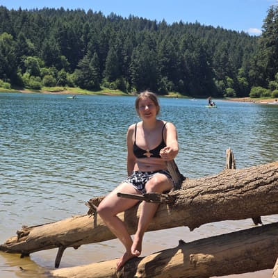 A young woman with blonde hair is sitting on a fallen tree trunk in the water at Hagg Lake, Oregon. She is wearing a black bikini top and black and white patterned shorts. The lake is calm and reflects the blue sky. A dense forest of green trees covers the hillside in the background. Other people can be seen in the distance on paddleboards and kayaks. The sun is shining brightly, casting shadows on the water.