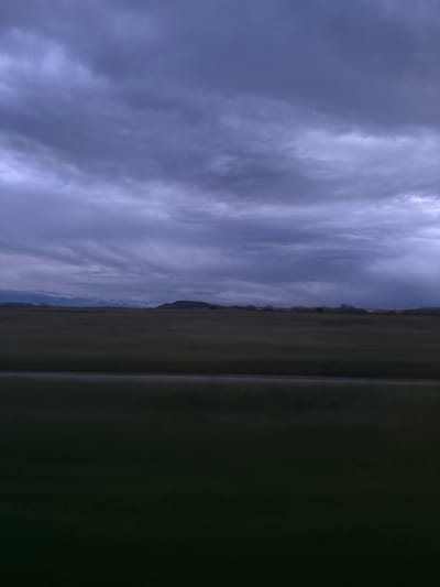 The image depicts a landscape in Tsar Asen, Bulgaria, under a cloudy sky. The left portion of the image showcases a dynamic, overcast sky filled with layers of gray clouds. To the right, the land is divided into two prominent sections: a darker, possibly waterlogged or shadowed area in the foreground and a strip of grassland running along the horizon. The grassland appears to be sparsely populated with trees. The horizon line is low and appears to separate the land from the sky. A few mountain peaks are discernible near the middle of the image.