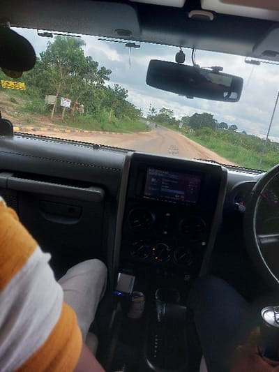 The image captures a scene from inside a dark-colored vehicle, likely a utility vehicle, looking out onto a dirt road in Kisanga, Tanzania. The foreground shows parts of two occupants: a person in a yellow and white striped shirt and light-colored pants occupies the passenger seat on the left, while a driver with dark pants and a hand on the steering wheel is visible on the right. The vehicle's dashboard features a central infotainment system displaying active content, climate control knobs with some illuminated indicators, an automatic transmission gear selector, and a cupholder containing a metallic can and a smaller cylindrical object. A rearview mirror hangs from the windshield, reflecting parts of the vehicle's interior.
The exterior view reveals a reddish-brown unpaved road that curves slightly to the right in the distance. The left edge of the road has a curb marked with black and yellow stripes. Lush green trees and dense vegetation flank both sides of the road. On the left side, a small white sign and a yellow sign are partially visible amidst the foliage. In the far distance, a small light-colored vehicle or motorcycle is present on the road. The sky is overcast with a mix of grey and white clouds.