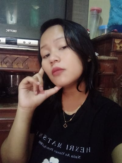 A woman poses for a photo with her hand to her chin, wearing a black t-shirt with text reading "HENRI MATISSE." She has short, dark hair and is wearing a gold necklace with a clover pendant. Behind her is a dark wooden cabinet with a television on top. Plastic containers are visible on the cabinet.
