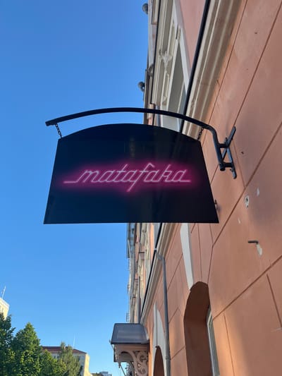 An outdoor, eye-level shot captures a neon sign spelling "matafaka" in pink, hanging from a building facade. The sign is black and mounted on a curved metal bracket. The building is painted a salmon pink and features architectural detailing. The sky is a clear blue. The setting is the city of Oulu, Finland.