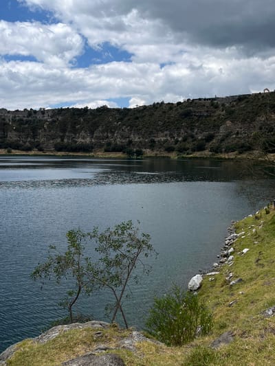 The image shows a panoramic view of the Aljojuca Lagoon in Aljojuca, Mexico. The lagoon is vast and dark blue, with ripples on its surface. The surrounding landscape is rugged and dry, with steep hills covered in scrubby vegetation. The sky is cloudy, with patches of blue visible. In the foreground, there are some rocks and grass, with a few small trees growing near the edge of the water. On the distant hills, there appear to be some structures, possibly buildings or observation points, and a small archway structure. The overall scene conveys a sense of natural beauty and remoteness.