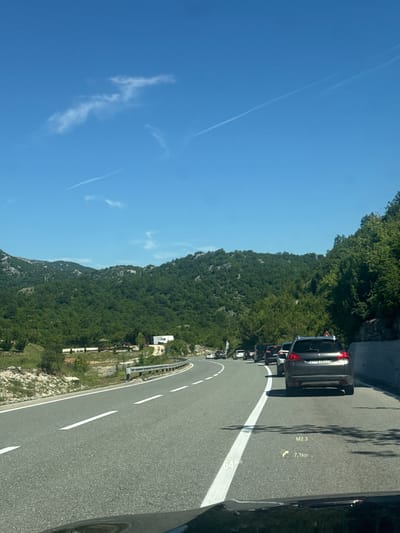 The image captures a scenic view of a winding road with a queue of cars on a sunny day, likely in Obzovica, Montenegro, given the context. The road is asphalt with clear white lane markings. Green, tree-covered mountains dominate the background, suggesting a mountainous or hilly terrain. The sky is a vibrant blue with a few wispy white clouds and a contrail. In the foreground, the dashboard of a vehicle is visible, displaying a heads-up display (HUD) with information such as "64 km/h," "M2.3," and "7,1km," indicating the vehicle's speed and distance to a point. Several cars are in traffic, moving along the road. One car in the immediate foreground is a dark-colored SUV with a license plate that appears to be "ZI-AJB23". On the left side of the road, there are some grassy slopes and vegetation. Further down the road, a small white building and a gazebo are visible amidst the greenery. The overall scene conveys a sense of travel and a picturesque mountainous landscape.