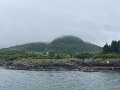 This image shows a rocky coastline with lush green vegetation leading up to a mountain shrouded in fog. Several buildings are visible nestled into the hillside, along with a Norwegian flag on a pole. The water in the foreground is choppy under an overcast sky. A red and white sign with an anchor symbol is visible on the rocks in the lower right corner of the image. The location is identified as Demmahågen, Norway.