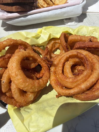 A plate of onion rings is on a table, some are in a yellow paper container. The onion rings are golden brown and appear to be deep-fried. There is also a sandwich and chips in the background. The scene is outdoors in what appears to be a restaurant patio. The location is Cannon Beach, United States of America.