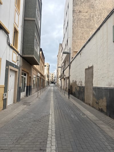 A street view in Arrecife, Spain, depicts a narrow roadway paved with gray brick tiles. The street is flanked by buildings of varying heights and architectural styles. The buildings are predominantly white and gray, with some having beige or yellow accents. The sky is overcast. The street appears deserted.