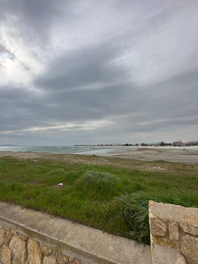 The image presents a wide coastal landscape in Limenas Avdiron, Greece, under an overcast sky. The foreground features a low wall composed of rough-hewn light brown stones, topped with a grey concrete slab. Directly beyond this wall, a strip of vibrant green grass, visibly wind-swept, extends towards the coastline. Within the grass, a small white object is distinguishable. This grassy area transitions into a broad, light-colored beach or intertidal zone, characterized by a mix of sand or mud, with scattered darker patches and white debris. The sea occupies the middle ground, displaying light blue-green water with moderate waves breaking towards the shore. Along the distant coastline, a horizontal line of buildings, primarily light-colored with some visible red roofs, indicates a settlement. A faint landmass is discernible on the far left horizon. The sky is heavily clouded with varying shades of grey, and brighter illumination originates from the upper left quadrant. No individuals or active subjects are present in the scene.