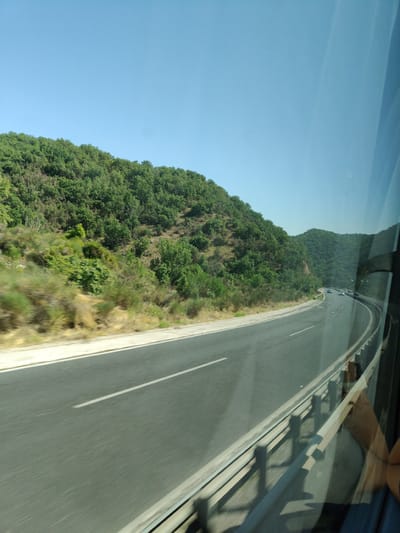 The image shows a view from a moving vehicle traveling on a highway. The highway curves to the right, flanked by a tree-covered hillside on the left and a guardrail on the right. The sky is clear and blue. Several vehicles are visible on the highway in the distance. The overall scene suggests a rural or semi-rural environment. While the prompt mentions the city of Rizana, Greece, there are no specific landmarks or features in the image that definitively place it in Rizana.