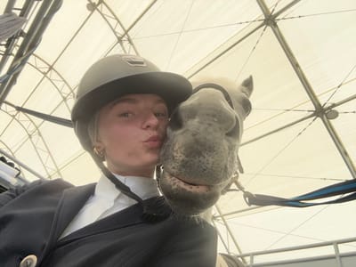 A young adult individual and a light-colored horse are depicted in an indoor equestrian facility, likely located in the Township of Langley, Canada. A female, positioned on the left, wears a dark equestrian helmet, a white collared shirt, and a dark riding jacket with a visible button. Her lips are pursed in a kissing gesture, directed towards the horse's head, which occupies the right side of the frame. The horse wears a dark bridle, and a blue and black lead rope is partially visible on its right side. The background reveals a large, translucent, light-colored fabric roof structure supported by an intricate network of metal trusses and cables, indicative of a covered riding arena.