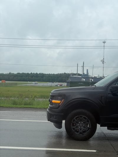 The image shows the side of a dark-colored vehicle, likely a truck or SUV, with its front orange turn signal illuminated. The vehicle is parked on a wet asphalt road with white lane markings, suggesting it might be raining or has recently rained. The environment appears to be an open area with a large, flat, grassy field to the left of the road. In the distance, behind the field, a line of trees forms a dark green backdrop. Further in the background, there are industrial structures, including tall smokestacks or chimneys and what appear to be shipping containers. There are also power lines stretching across the sky, and a distant transmission tower is visible. The sky is overcast with clouds, indicating a cloudy or gloomy day. The image is taken from a low angle, focusing on the vehicle and the immediate surroundings. There is no visible text in the image. The overall mood is neutral and observational.