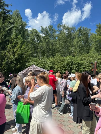 The image shows an outdoor gathering of people, likely at a community event or festival, under a clear blue sky with scattered clouds and surrounded by lush green trees. In the foreground and midground, a diverse group of individuals, predominantly women of various ages, are seen standing and interacting. Many are holding disposable bowls and cups, suggesting they are being served food or drinks. Some individuals are actively eating or being served. The setting appears to be a paved area, possibly a park or a public square, with a large tent or canopy structure in the background, which could be used for catering or shelter.

A person wearing a red shirt with "СПАСАТЕЛЬ" (Rescuer) printed on the back is visible, suggesting some form of organized activity or event. A bright green reusable shopping bag is held by one of the women, adding a splash of color to the scene. The overall atmosphere suggests a casual, social event taking place on a sunny day. The specific location in Duvankul', Russia, is not explicitly visible in the image, but the presence of a diverse crowd and outdoor setting are consistent with a local event in such a location.