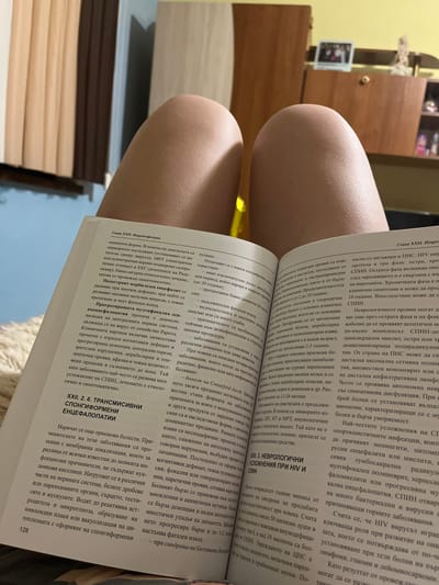 The image shows an open book with Bulgarian text. The book is placed on a person's lap, as evidenced by the smooth, pale skin of a leg and knee visible in the foreground and partially obscuring the lower left corner of the book. The background consists of a slightly out-of-focus wooden piece of furniture with some items on top, possibly in a room. The text on the pages discusses medical topics, with headings such as "Глава XXII. Невроинфекции" (Chapter XXII. Neuroinfections) and "XXII. 2. 6. ТРАНСМИСИВНИ СПОНГІФОРМЕНИ ЕНЦЕФАЛОПАТИИ" (XXII. 2. 6. TRANSMISSIBLE SPONGIFORM ENCEPHALOPATHIES). The lighting suggests an indoor setting, likely during the daytime or with artificial light. The book appears to be a medical textbook or reference. There is no information or visual cues within the image that would indicate the location in the city of Popintsi, Bulgaria.