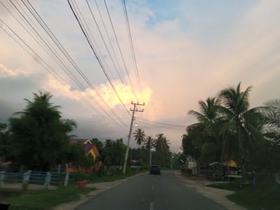 A paved road runs through a rural area, with a dark-colored car driving away from the camera. The road is bordered by vegetation and a few houses, one of which is yellow with a purple roof. Overhead, a network of power lines crisscrosses the sky, which is filled with clouds and a soft, warm glow suggesting sunset or sunrise. Several palm trees are visible on either side of the road. The scene appears to be set in a tropical or subtropical location, likely in or near Meuse, Indonesia, given the presence of palm trees and the architectural style of the houses. The lighting and atmosphere suggest a peaceful, twilight moment.