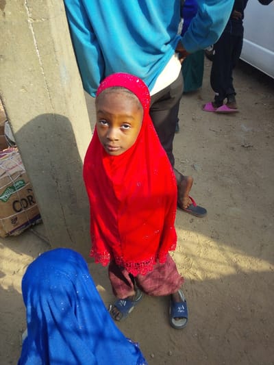 In Unguwa Uku Kauyen Alu, Nigeria, a young child with dark skin, wearing a bright red hijab with lace trim and patterned maroon trousers, stands prominently in the foreground, looking directly at the camera with a neutral expression. The child wears blue-and-white slide sandals. Directly behind the child, the lower torso and legs of an adult wearing a light blue long-sleeved shirt and dark trousers are visible, with their right hand placed on their hip. To the immediate left of the child is a weathered concrete pillar. Behind the pillar, stacked cardboard boxes, some labeled "WHOLE GRAIN" and "CORN," are visible. In the bottom-left foreground, another individual is partially visible, wearing a royal blue head covering with small reflective elements, appearing to be seated or kneeling. In the background, other individuals are present, including one wearing dark trousers and pink flip-flops, and a portion of a white vehicle. The ground is packed dirt or sand, exhibiting areas of both direct sunlight and shadow, suggesting an outdoor daytime setting.