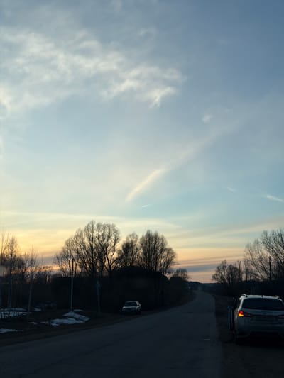 The image shows a partially cloudy sky with a road and trees to the right, a car on the road, and a patch of snow beside it. The setting appears to be at dusk or dawn given the color of the sky. Location could not be identified based on image alone.