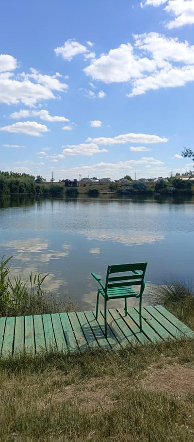 The image depicts a serene lakeside scene in the village of Vilinskoye, Ukraine. A bright green, metal chair stands on a weathered wooden dock, facing a calm body of water. The lake reflects the clear blue sky, dotted with fluffy white clouds. The shoreline on the opposite side of the lake features a line of trees and a few scattered buildings, including some greenhouses. Tall grasses and reeds grow along the edge of the dock, adding a touch of wildness to the scene. The overall impression is one of peace and tranquility, a perfect spot for quiet contemplation by the water.