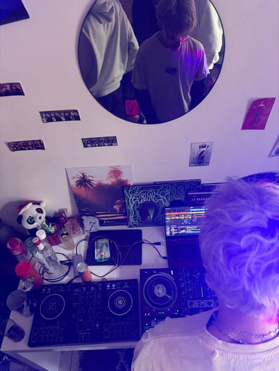 This image depicts an indoor scene in Saint-Leu-la-Forêt, France, viewed from an over-the-shoulder perspective of an individual engaged with electronic music equipment. A person with short, light-colored, wavy hair, wearing a light-colored t-shirt and multiple thin necklaces, is positioned with their back to the viewer, standing before a white desk.
On the desk, a black Pioneer DJ controller is centrally placed. To its right, an open laptop displays DJ software with colorful audio waveforms and a mixer interface. A small black rectangular device, likely an external hard drive or audio interface, is situated between the laptop and the controller, connected by cables. The left side of the desk is cluttered with objects including a white panda plush toy wearing a red Santa hat, several empty and partially filled clear glasses and plastic cups, a clear glass bottle, two aluminum cans (one silver, one orange), a gray power adapter, and a small white device with a colorful screen.
The white wall above the desk features a large oval mirror reflecting at least two individuals. One reflection shows a person resembling the foreground subject, wearing a light t-shirt and glasses, with a prominent purple light illuminating their chest. Another reflection reveals the torso of a person in a white hoodie to the left of the first reflection. The wall is adorned with four strips of photo booth-style pictures. Below these, a poster depicts a tropical sunset or sunrise scene with palm trees, labeled "BALI INDONESIA." To its right, two square artworks display intricate organic green designs, one clearly labeled "BIAR." Further right, two smaller posters are affixed: one with a red artistic rendering of a figure, and another featuring "SELENA GOMEZ."
The scene is uniformly illuminated by a strong purple-blue artificial light source, creating a distinct ambiance.