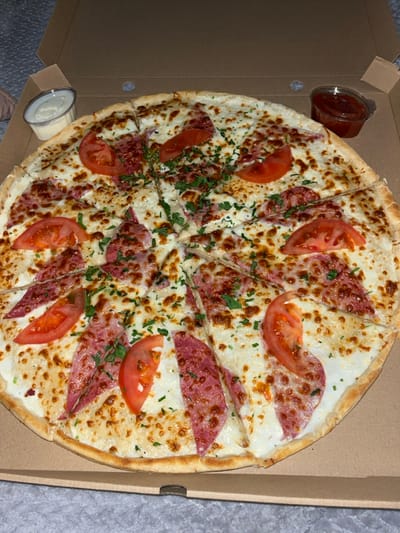 A large, round pizza, segmented into eight slices, rests within an open brown cardboard delivery box. The pizza features a white sauce base, generously covered with melted, lightly browned cheese. Distributed across the pizza are multiple crescent-shaped slices of a red cured sausage, approximately ten slices of red tomato, and finely chopped green herbs. Two small, clear plastic condiment cups are positioned in the top corners of the box; one contains a white creamy substance, and the other holds a red sauce. The pizza box is situated on a gray, textured fabric surface. No individuals are visible. This scene is located in the city of Zlatovrah, Bulgaria.