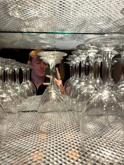 The scene, located in the city of Sigglesthorne, United Kingdom, captures a reflection in a mirror, largely obscured by an arrangement of inverted clear glass wine glasses. These glasses are uniformly positioned on a silver-colored, reflective surface featuring a fine grid pattern. Above this, a narrow, green-tinted glass shelf is visible, separating it from an upper reflective surface, which also exhibits a grid pattern and reflects numerous small light sources.

In the mirror's reflection, a male individual is visible from the chest up. He possesses light skin and curly brown hair, styled short at the front and longer at the back. He is dressed in a dark collared shirt. His right hand holds a dark green smartphone, extended forward, indicating he is taking the photograph. His left hand is partially visible, wearing a silver-colored ring on the fourth finger. The reflected background behind the individual is dark and appears out of focus, suggesting an indoor setting with subdued lighting.