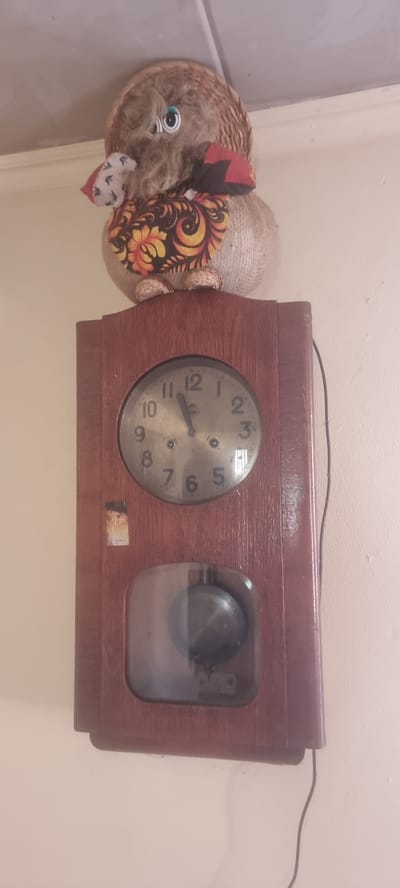 This is a photograph of a wooden mantel clock and a decorative object resembling a pig. The clock is made of dark wood and has a circular face with Arabic numerals and black hands. The time displayed is approximately 9:49. To the left of the clock face, there is a rectangular glass-covered compartment showing a pendulum swinging. The decorative object next to the clock appears to be a pig-shaped figurine with a woven straw body, a tuft of grey hair for a snout, and large googly eyes. It is adorned with colorful, floral fabric, possibly in a traditional Russian folk art style, and red and black fabric accents on its ears. The objects are placed against a plain, light-colored wall. A dark electrical cord runs from the top of the clock towards the right side of the frame. The lighting suggests an indoor setting, possibly during the day. No visible text is present on the clock or the figurine. The setting cues, such as the decorative fabric style, align with the provided location context of Chernomorskiy, Russia.